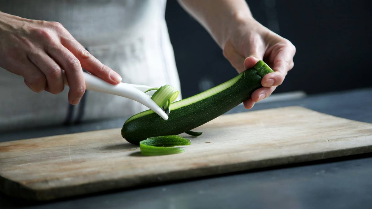Comparison of four peelers including a stainless steel potato peeler next to peeled vegetables on wooden counter